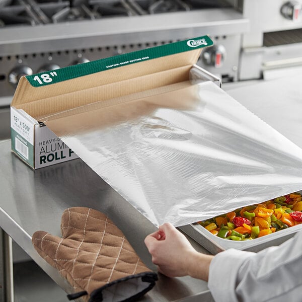 A person's hand unrolling Choice heavy-duty aluminum foil over a tray of food.